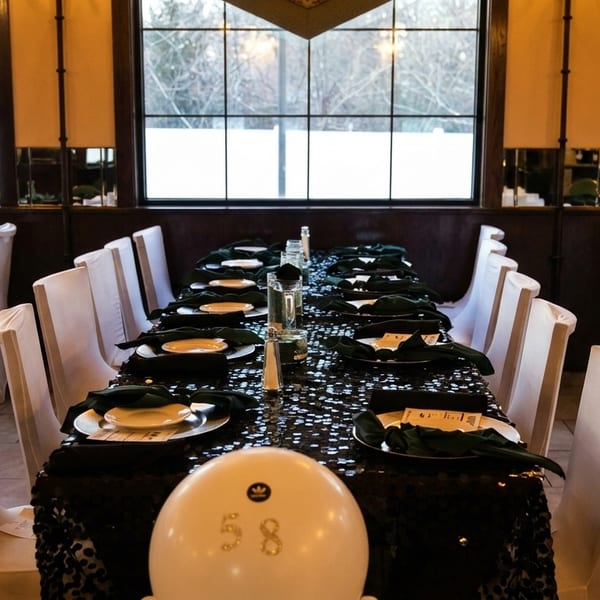 A long dining table set with dark tablecloths, white plates, and green napkins, with white chairs.