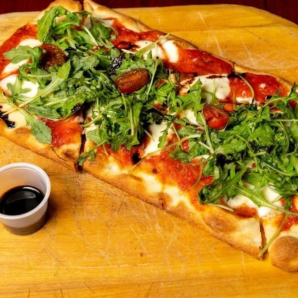 A rectangular flatbread pizza with tomato sauce, mozzarella, arugula, and cherry tomatoes on a wooden peel.