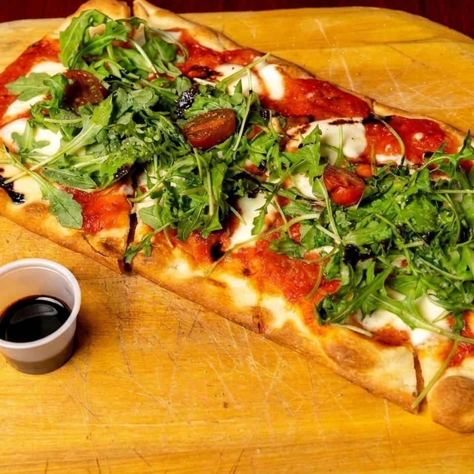 A rectangular flatbread pizza with tomato sauce, mozzarella, arugula, and cherry tomatoes on a wooden peel.