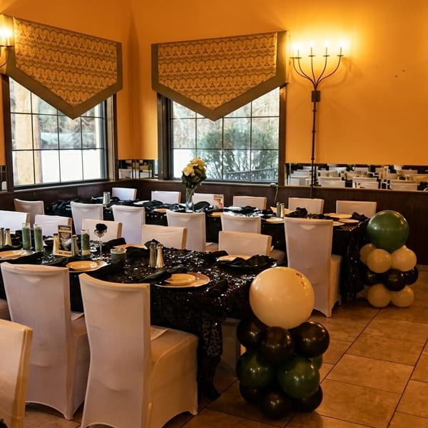 A restaurant dining room set for an event with black sequin tablecloths, white chairs, and balloon decorations.