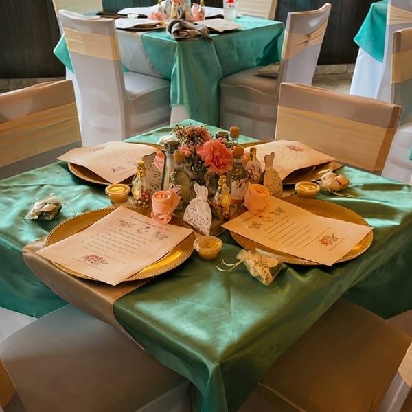 Restaurant dining room with tables draped in teal cloths, set with gold chargers, menus, and floral centerpieces.