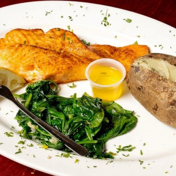 Grilled salmon with a lemon wedge, sautéed spinach, a baked potato, and melted butter on a white plate.
