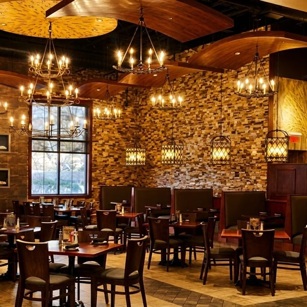 Warmly lit restaurant interior with dark wood tables, chairs, and booths, stone walls, and ornate chandeliers.